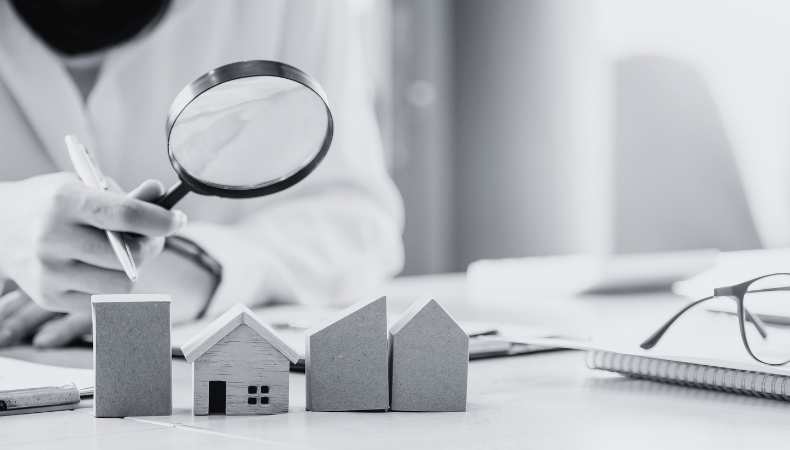 A black and white image of a person holding a magnifying glass looking over a number of miniature sized properties placed on a table.