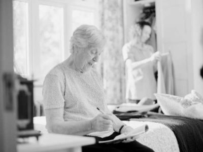 A black and white image of an older lady on a bed writing notes in an assisted living property.