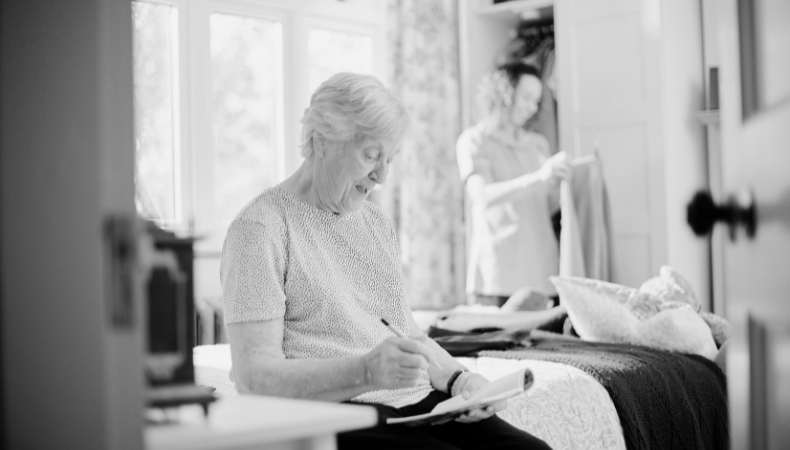 A black and white image of an older lady on a bed writing notes in an assisted living property.