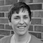 Black and white headshot of woman with short hair smiling with a brick wall in the background.