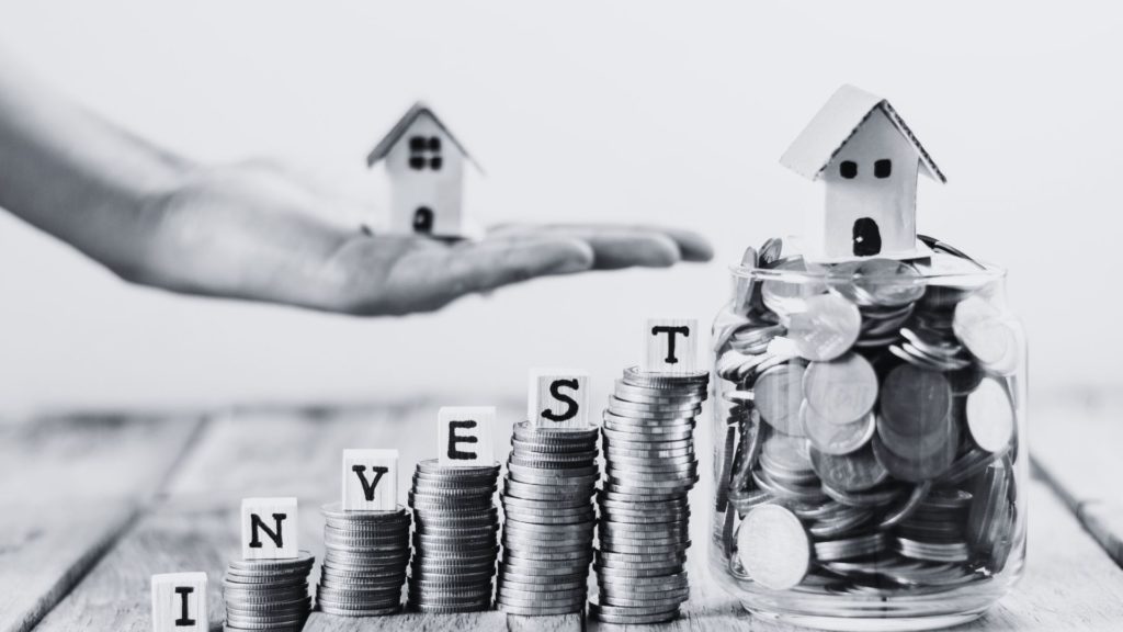 Black and white coin towers with invest in dice letters with the word invest featuring a hand with a model house.