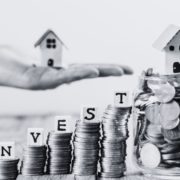 Black and white coin towers with invest in dice letters with the word invest featuring a hand with a model house.