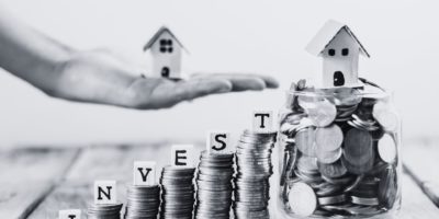 Black and white coin towers with invest in dice letters with the word invest featuring a hand with a model house.