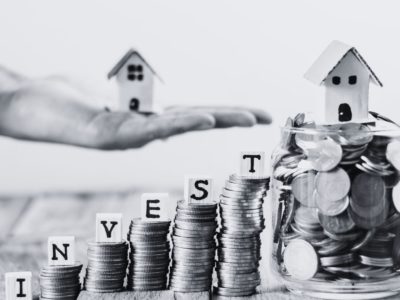 Black and white coin towers with invest in dice letters with the word invest featuring a hand with a model house.