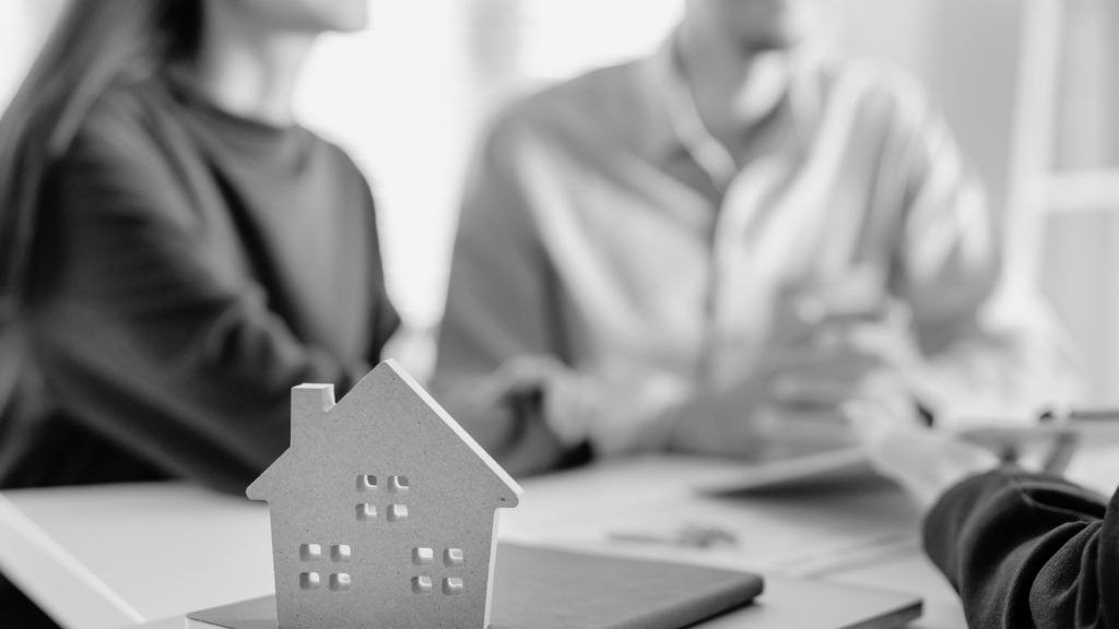 Two people talking to another person other a desk with a cut out house.